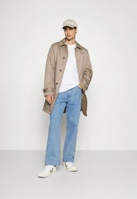 Šviesiai rudos spalvos lengvas tranšėjos paltas, balti marškinėliai, šviesiai mėlyni džinsai ir balti sportbačiai, su šviesiai rudos spalvos kepure ant modelio prieš paprastą foną.