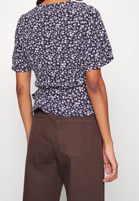 Blusa floreale blu navy con maniche corte, caratterizzata da una vita stretta e un orlo a peplo. Abbinata a pantaloni marroni a vita alta visti da dietro.