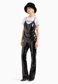 Mono de lentejuelas negras con escote drapeado, combinado con una camiseta gráfica blanca y un sombrero de cubo de piel sintética negra. Un calzado brillante completa el look.