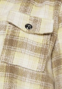 Skjorte med plaid-mønster, der har en blød og fuzzy tekstur i beige, creme og gul med en sort knap på brystlommen og synlige broderidetaljer.