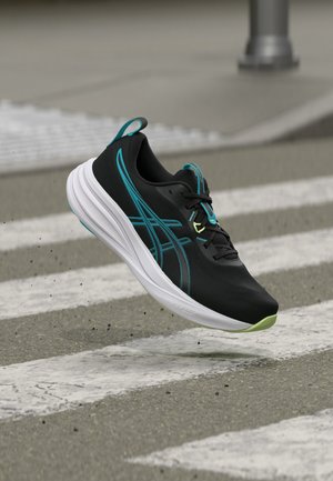 Zapatilla de correr negra con detalles en azul verdoso y suela blanca suspendida sobre un paso de peatones, levantando pequeños escombros.
