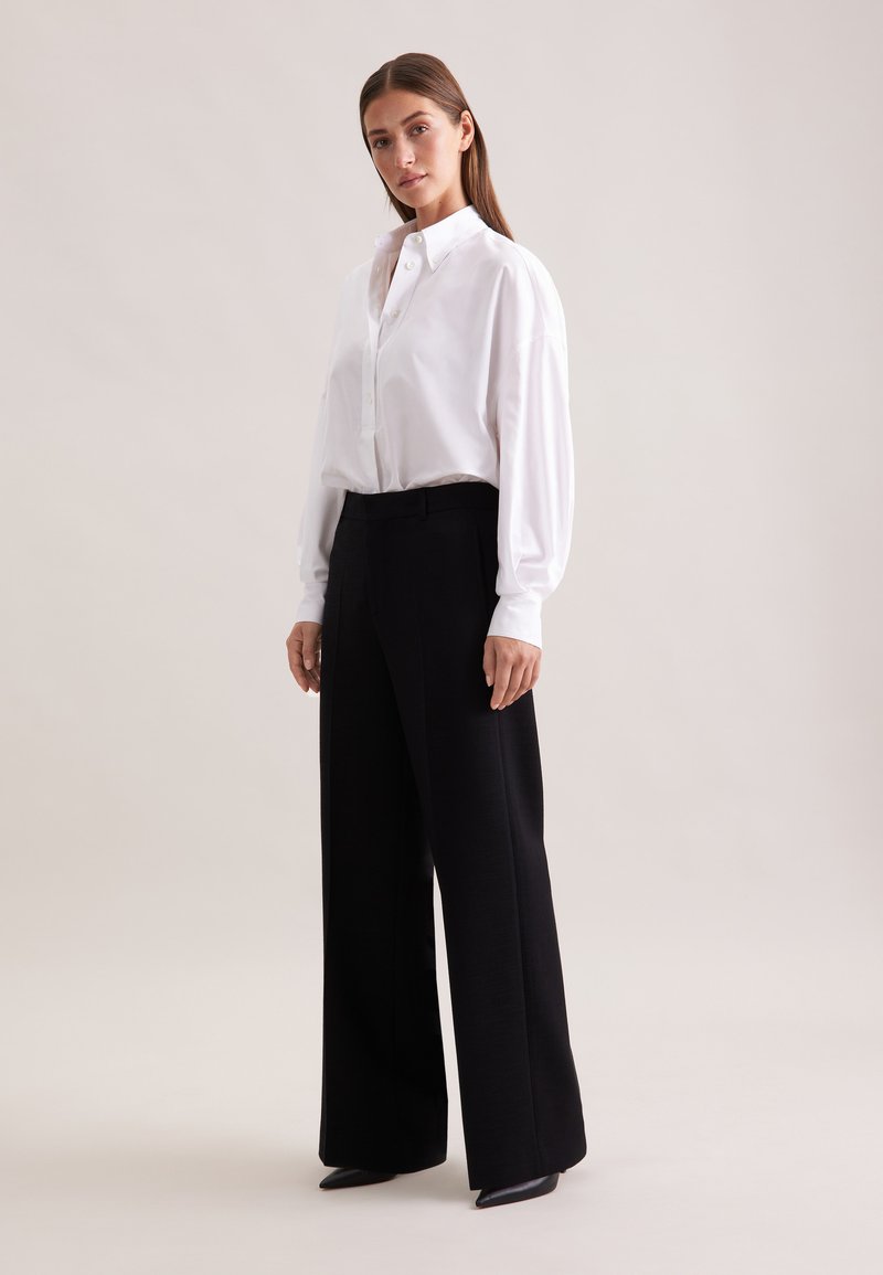 Camicia bianca con bottoni, maniche lunghe, abbinata a pantaloni neri a vita alta e gamba larga, e scarpe nere con punta su uno sfondo neutro.