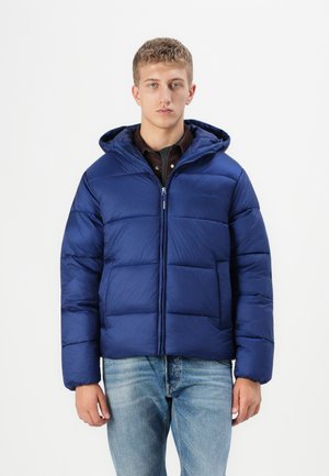 Marineblauwe gewatteerde jas met een capuchon, ritssluiting en gestikte panelen. Voorzien van een voorzak en een textuurstof. Draagt over een donker shirt.