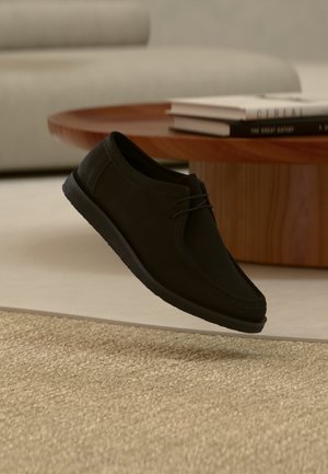 Zwarte leren veterschoen zwevend boven beige gestructureerd tapijt, met een houten ronde tafel en gestapelde boeken op de zacht gefocuste achtergrond.