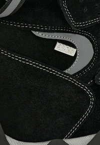 Schwarze Wildleder-Schuh mit grauen Akzenten, weißen Nähten und einem wasserdichten Etikett. Das Design umfasst eine geschwungene Form und eine strukturierte Oberfläche.