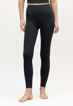 Frau trägt schwarze Leggings mit hohem Bund und ein schwarzes, kurzes Top, steht barfuß vor einem schlichten hellen Hintergrund.