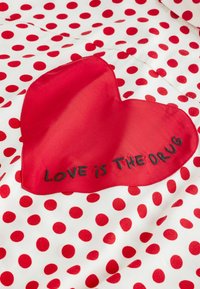 Roter Herzaufnäher mit dem Schriftzug "LOVE is THE DRUG", auf weißem Stoff mit roten Punkten aufgenäht.