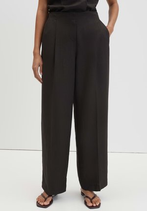 Person wearing high-waist, loose black wide-leg trousers with side pockets paired with black thong sandals, standing on light floor.