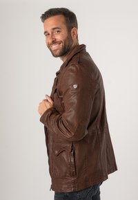 Braune Lederjacke mit strukturiertem Finish, ausgestattet mit einem Reißverschluss, seitlichen Taschen und einem Wappen-Logo auf der linken Schulter.