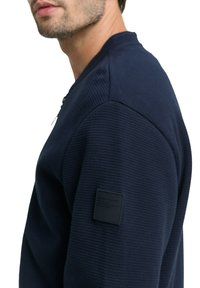 Granatowa teksturowana bluza z prążkowanym wzorem, z czarną etykietą na lewym rękawie i okrągłym dekoltem.