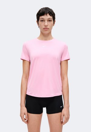 Žena s kratkom tamnom kosom u svijetloružičastoj Nike majici i crnim Nike kratkim hlačama stoji na jednostavnoj svijetloj pozadini.