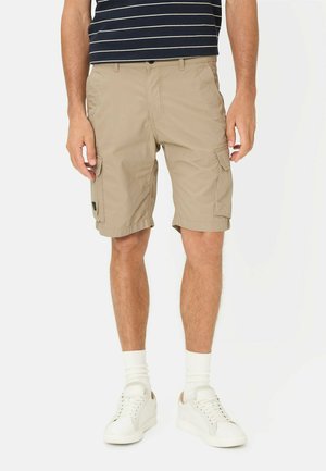 Man draagt beige cargoshorts, witte sneakers en witte sokken, staat tegen een effen witte achtergrond.
