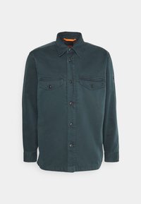 Chemise à manches longues vert foncé avec deux poches poitrine boutonnées et un col à pointes étalées sur fond blanc.