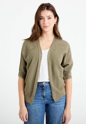 Joven con cabello largo y castaño que lleva pendientes de aro dorados, un cárdigan abierto de textura oliva clara, camiseta blanca y vaqueros azules, de pie.