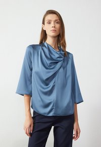 Mėlyna satino blusa su drapiruotu iškirpimu, trijų ketvirčių rankovėmis ir laisvu siluetu, derinama su tamsiomis kelnėmis. Glotni tekstūra, minimalistinis dizainas.