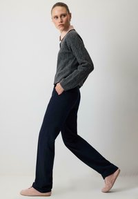 Pulover gri tricotat, pantaloni drepți bleu închis și adidași roz deschis. Model mergând cu mâinile în buzunare, fundal neutru.