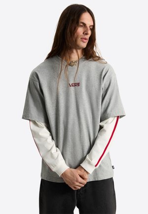 Persona con i capelli lunghi che indossa una maglietta grigia Vans sopra una camicia bianca a maniche lunghe con strisce rosse, in piedi davanti a uno sfondo semplice.