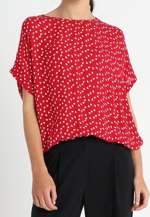 Femme portant une blouse rouge ample avec des motifs de cœurs blancs et un pantalon noir taille haute, debout devant un fond blanc uni.