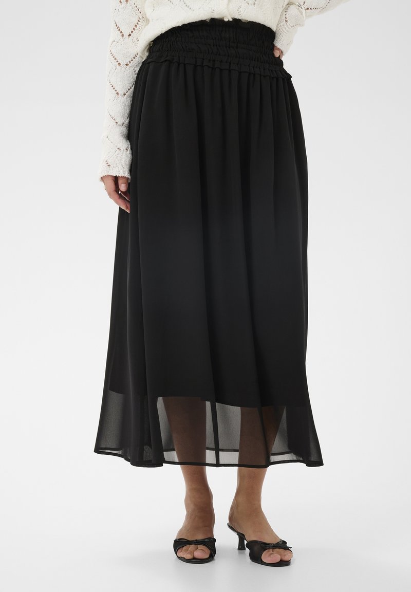 Femme portant une jupe midi noire à ourlet transparent, un pull en maille blanc et des sandales à talons ouvertes noires, debout devant un fond uni.