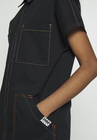 Chemise noire à manches courtes avec des coutures contrastantes en rose, orange et bleu. Comprend deux poches poitrine et une étiquette sur le côté.