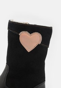 Botín negro de ante hasta el tobillo con una sección inferior de cuero negro suave. Presenta un parche en forma de corazón rosa con acentos brillantes en la parte superior.