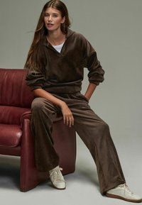 Hnědý velurový teplákový komplet s polovičním zipem a kalhotami s širokými nohavicemi, zkombinovaný s bílými teniskami. Texturovaná látka s volným střihem.