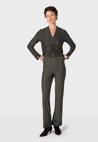 Femme debout, mains sur les hanches, portant un haut à manches longues assorti noir et blanc à motifs et un pantalon évasé avec des chaussures à talons noires.