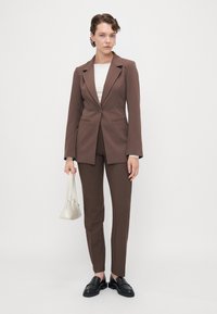 Brun skreddersydd blazer og matchende bukser, kombinert med en hvit topp, svarte loafers og en liten hvit håndveske. Glatt stoff med strukturert design.