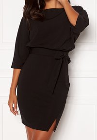 Abito nero con scollatura drappeggiata, maniche a tre quarti, vita con cintura, silhouette aderente e uno spacco alto fino alla coscia su un lato.