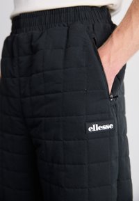 Jogging en tissu matelassé noir avec une taille élastique, une poche zippée sur le côté et un grand logo "ellesse" en blanc sur la cuisse.