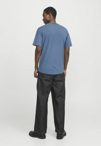 Blue short-sleeve t-shirt with a curved hem, paired with wide black denim pants and black shoes, viewed from the back.