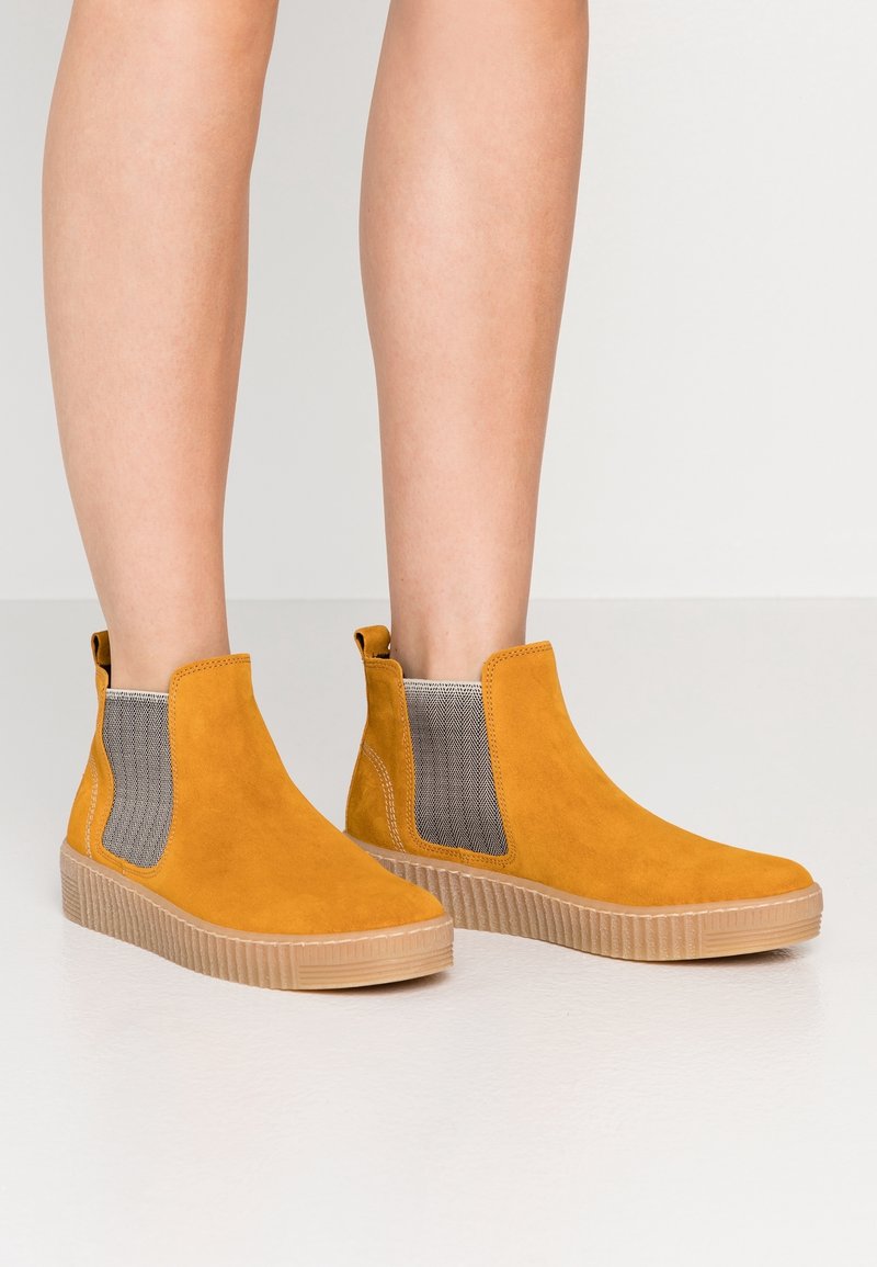 Jambes portant des bottines Chelsea en daim jaune moutarde avec des panneaux élastiques latéraux beiges et des semelles épaisses en caoutchouc sur un fond clair uni.