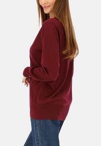 Bordowy sweter z dzianiny z długimi rękawami, okrągłym dekoltem oraz prążkowanymi ściągaczami na mankietach i w dole. W połączeniu z niebieskimi dżinsami o luźnym kroju.
