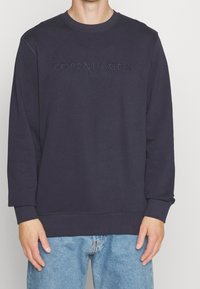 Sudadera azul marino hecha de una suave mezcla de algodón, con cuello redondo y texto en relieve "COPENHAGUE" en la parte delantera, combinada con jeans de color azul claro.