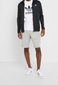 Černá atletická bunda na zip s bílými pruhy, bílý tričko s černým logem, šedé šortky a bílé tenisky s barevnými akcenty.