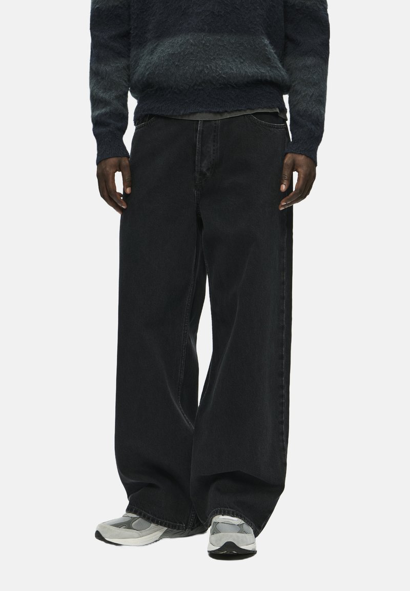 Zwart wijde jeans met een klassiek vijfzakkenontwerp, zware denimstof en subtiele stikdetails, gecombineerd met grijze sneakers.