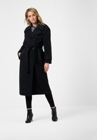 Svart trenchcoat i mjukt tyg, dubbelknäppt design med breda slag, bälte i midjan och knäppte ärmar. Stylad med svarta byxor och stövlar.