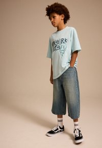 T-shirt graphique bleu clair avec impression noire, associée à un short en denim ample, des baskets noires avec des touches blanches et des chaussettes rayées.