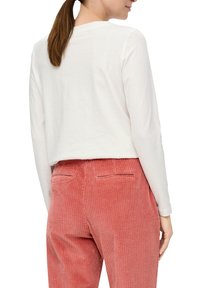Kobieta z brązowym kucykiem, ubrana w białą bluzkę z długim rękawem oraz różowe spodnie sztruksowe z wysokim stanem, stoi tyłem.