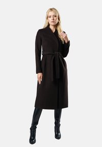 Kobieta stojąca w długim czarnym płaszczu z paskiem i czarnych botkach na wysokim obcasie na jednolitym białym tle.