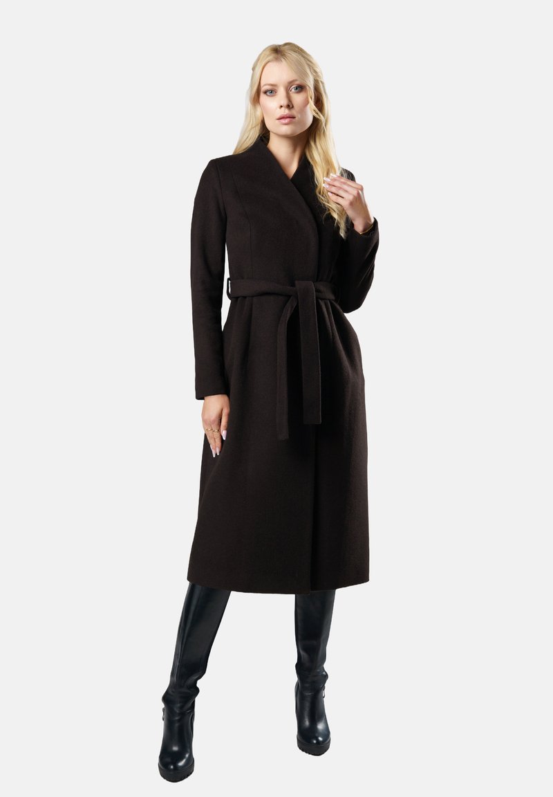 Kobieta stojąca w długim czarnym płaszczu z paskiem i czarnych botkach na wysokim obcasie na jednolitym białym tle.