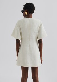 Robe mini blanche texturée avec des manches courtes, dotée d'une coupe droite, d'une couture au dos et d'un décolleté épuré. Le modèle porte de grandes boucles d'oreilles en or.