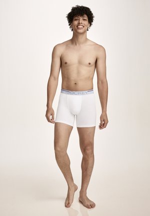 Lächender Mann steht barfuß und trägt weiße Boxershorts mit gebrandetem Bund vor einem schlichten hellen Hintergrund.