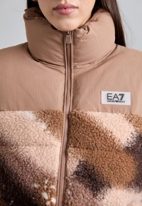 Ženska, oblečena v bež in rjavo jakno z visokim ovratnikom na zadrgo, ki ima mešan dizajn tkanin in vidno oznako EA7 Emporio Armani.