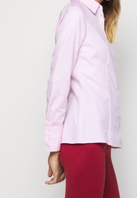 Camicia button-up rosa chiaro con colletto e maniche lunghe, dotata di bottoni rotondi lungo il fronte e i polsini. Tessuto dalla consistenza liscia.