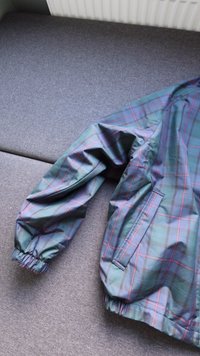 Casaco corta-vento em padrão xadrez verde e azul, com punhos elásticos, um bolso lateral e tecido leve e suave.