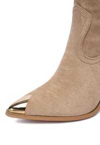 Bézs színű velúrbőr bokacsizma hegyes orral, amely fényes aranyszínű orrlemezt kapott, sima felülettel, karcsú, minimalista kialakítással.