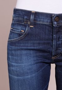 Dżinsy jeansowe w ciemnoniebieskim kolorze z delikatną fakturą, wyposażone w pięć kieszeni, kontrastowe przeszycia oraz metalowe zapięcie na guzik w pasie.