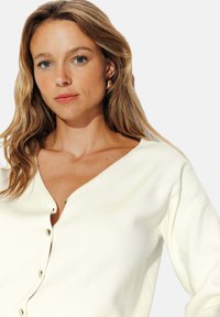 Le cardigan côtelé de couleur crème présente un col en V, une fermeture à boutons et des manches longues. Le tissu a une texture douce avec un minimum de détails.