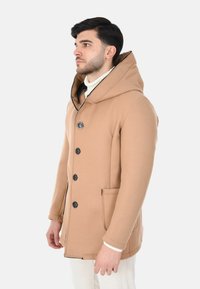 Cappotto con cappuccio color cammello con bottoni neri, tasche laterali e dettagli di cuciture visibili. Tessuto liscio con una vestibilità strutturata, indossato sopra un maglione bianco.